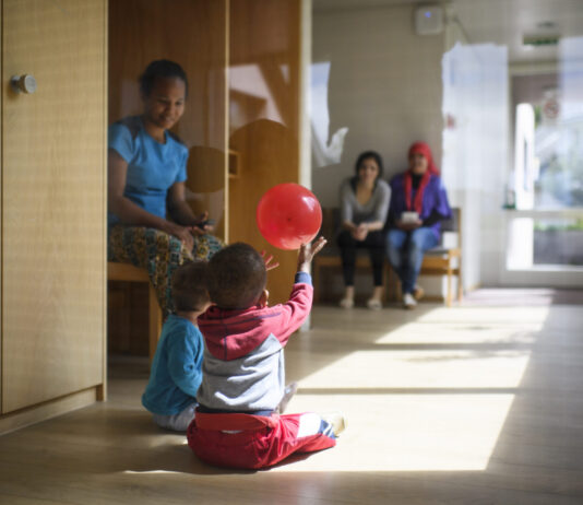 L’UDC veut démanteler le droit d’asile — les conséquences pour les réfugié-es sont dramatiques Dans un couloir lumineux d'un centre d'asile en Suisse alémanique au sol en bois, deux tout-petits sont assis par terre ; l’un d’eux lève les bras pour attraper un ballon rouge qui flotte au-dessus de sa tête. À gauche, une femme est assise sur un banc, les observant, et en arrière-plan deux autres adultes sont également assis, tournés vers les enfants.