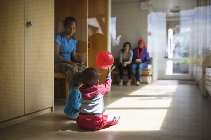 ASYLWESEN, MIGRATION, ASYLHEIM, ASYLUNTERKUNFT, ASYLZENTRUM, Dans un couloir lumineux d'un centre d'asile en Suisse alémanique au sol en bois, deux tout-petits sont assis par terre ; l’un d’eux lève les bras pour attraper un ballon rouge qui flotte au-dessus de sa tête. À gauche, une femme est assise sur un banc, les observant, et en arrière-plan deux autres adultes sont également assis, tournés vers les enfants.