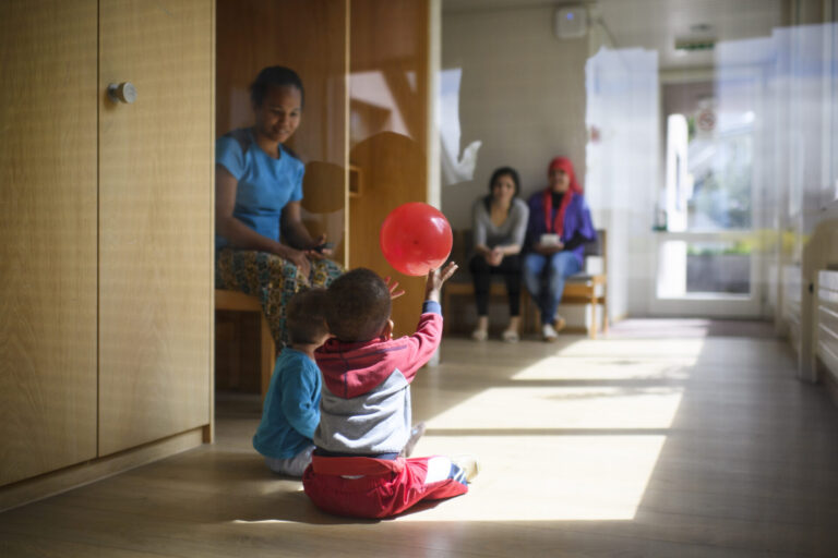 Dans un couloir lumineux d'un centre d'asile en Suisse alémanique au sol en bois, deux tout-petits sont assis par terre ; l’un d’eux lève les bras pour attraper un ballon rouge qui flotte au-dessus de sa tête. À gauche, une femme est assise sur un banc, les observant, et en arrière-plan deux autres adultes sont également assis, tournés vers les enfants.