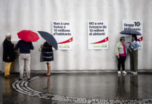 Initiative pour le chaos : voici comment l’UDC veut détruire la Suisse Photo prise sous la pluie devant un mur gris orné de trois affiches de l’UDC – à gauche en français : “Non à une Suisse à 10 millions d’habitants”, au centre en italien : “NO a una Svizzera da 10 milioni di abitanti”, à droite en allemand : “Stopp 10-Millionen…”. Au sol mouillé, on aperçoit le reflet des silhouettes. À gauche, un petit groupe de trois personnes debout sous leurs parapluies discute près d’une boîte posée contre le mur. À droite, deux femmes échangeant elles aussi sous un parapluie se font face.