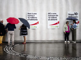 Initiative pour le chaos : la dangereuse expérimentation de l’UDC Photo prise sous la pluie devant un mur gris orné de trois affiches de l’UDC – à gauche en français : “Non à une Suisse à 10 millions d’habitants”, au centre en italien : “NO a una Svizzera da 10 milioni di abitanti”, à droite en allemand : “Stopp 10-Millionen…”. Au sol mouillé, on aperçoit le reflet des silhouettes. À gauche, un petit groupe de trois personnes debout sous leurs parapluies discute près d’une boîte posée contre le mur. À droite, deux femmes échangeant elles aussi sous un parapluie se font face.