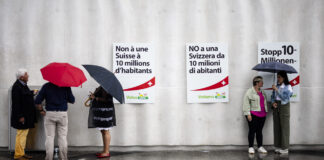 Initiative pour le chaos : la dangereuse expérimentation de l’UDC Photo prise sous la pluie devant un mur gris orné de trois affiches de l’UDC – à gauche en français : “Non à une Suisse à 10 millions d’habitants”, au centre en italien : “NO a una Svizzera da 10 milioni di abitanti”, à droite en allemand : “Stopp 10-Millionen…”. Au sol mouillé, on aperçoit le reflet des silhouettes. À gauche, un petit groupe de trois personnes debout sous leurs parapluies discute près d’une boîte posée contre le mur. À droite, deux femmes échangeant elles aussi sous un parapluie se font face.