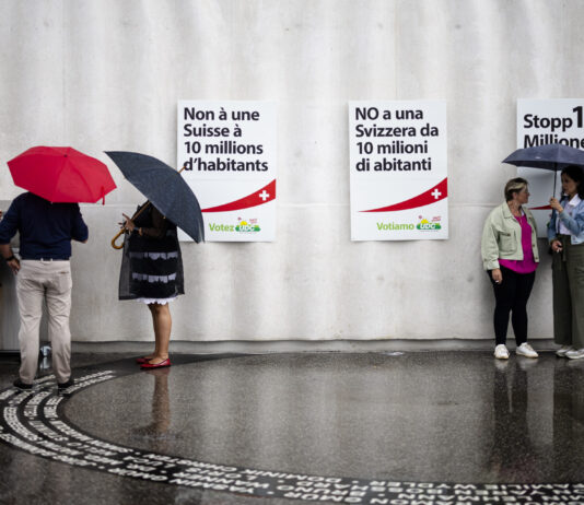 Initiative pour le chaos : la dangereuse expérimentation de l’UDC Photo prise sous la pluie devant un mur gris orné de trois affiches de l’UDC – à gauche en français : “Non à une Suisse à 10 millions d’habitants”, au centre en italien : “NO a una Svizzera da 10 milioni di abitanti”, à droite en allemand : “Stopp 10-Millionen…”. Au sol mouillé, on aperçoit le reflet des silhouettes. À gauche, un petit groupe de trois personnes debout sous leurs parapluies discute près d’une boîte posée contre le mur. À droite, deux femmes échangeant elles aussi sous un parapluie se font face.