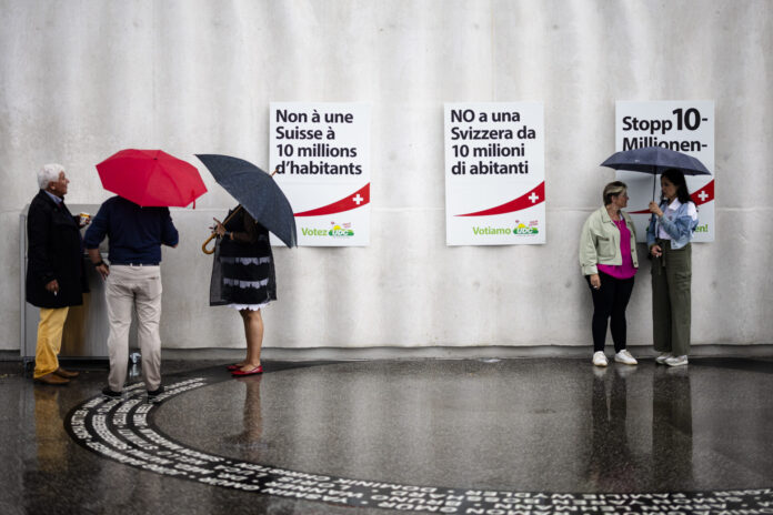 Photo prise sous la pluie devant un mur gris orné de trois affiches de l’UDC – à gauche en français : “Non à une Suisse à 10 millions d’habitants”, au centre en italien : “NO a una Svizzera da 10 milioni di abitanti”, à droite en allemand : “Stopp 10-Millionen…”. Au sol mouillé, on aperçoit le reflet des silhouettes. À gauche, un petit groupe de trois personnes debout sous leurs parapluies discute près d’une boîte posée contre le mur. À droite, deux femmes échangeant elles aussi sous un parapluie se font face.
