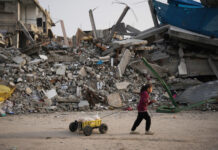 Palestine : Le Conseil fédéral doit faire plus Une fillette dans la bande de Gaza, en sweat à capuche bordeaux et pantalon sombre, avance sur un sol poussiéreux, tirant derrière elle une petite charrette artisanale — un bac plastique jaune sur quatre roues chargé de pains enveloppés de sacs — tandis qu’en arrière-plan s’étend un vaste tas de décombres : béton effondré, barres d’armature tordues et pièces métalliques brisées d’un bâtiment détruit.
