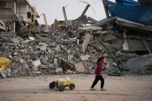 Une fillette dans la bande de Gaza, en sweat à capuche bordeaux et pantalon sombre, avance sur un sol poussiéreux, tirant derrière elle une petite charrette artisanale — un bac plastique jaune sur quatre roues chargé de pains enveloppés de sacs — tandis qu’en arrière-plan s’étend un vaste tas de décombres : béton effondré, barres d’armature tordues et pièces métalliques brisées d’un bâtiment détruit.