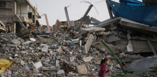 Palestine : le Conseil fédéral doit faire plus Une fillette dans la bande de Gaza, en sweat à capuche bordeaux et pantalon sombre, avance sur un sol poussiéreux, tirant derrière elle une petite charrette artisanale — un bac plastique jaune sur quatre roues chargé de pains enveloppés de sacs — tandis qu’en arrière-plan s’étend un vaste tas de décombres : béton effondré, barres d’armature tordues et pièces métalliques brisées d’un bâtiment détruit.