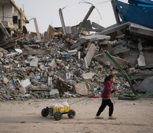 Palestine : le Conseil fédéral doit faire plus Une fillette dans la bande de Gaza, en sweat à capuche bordeaux et pantalon sombre, avance sur un sol poussiéreux, tirant derrière elle une petite charrette artisanale — un bac plastique jaune sur quatre roues chargé de pains enveloppés de sacs — tandis qu’en arrière-plan s’étend un vaste tas de décombres : béton effondré, barres d’armature tordues et pièces métalliques brisées d’un bâtiment détruit.