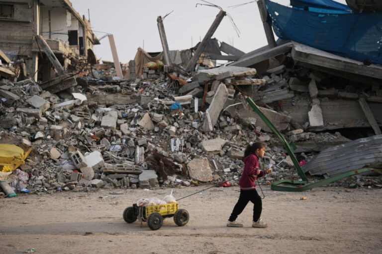 Une fillette dans la bande de Gaza, en sweat à capuche bordeaux et pantalon sombre, avance sur un sol poussiéreux, tirant derrière elle une petite charrette artisanale — un bac plastique jaune sur quatre roues chargé de pains enveloppés de sacs — tandis qu’en arrière-plan s’étend un vaste tas de décombres : béton effondré, barres d’armature tordues et pièces métalliques brisées d’un bâtiment détruit.
