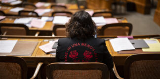 Un demi-million de signatures : quand les femmes se mobilisent Vue de dos de Tamara Funiciello, assise dans un hémicycle au mobilier en bois, des rangées de bureaux jonchés de dossiers, de feuilles et d’un ordinateur portable devant elle, portant une veste noire ornée en rouge de l’inscription “Ni Una Menos” entourant deux roses stylisées.