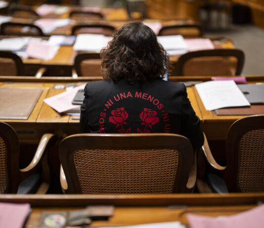 Un demi-million de signatures : quand les femmes se mobilisent Vue de dos de Tamara Funiciello, assise dans un hémicycle au mobilier en bois, des rangées de bureaux jonchés de dossiers, de feuilles et d’un ordinateur portable devant elle, portant une veste noire ornée en rouge de l’inscription “Ni Una Menos” entourant deux roses stylisées.