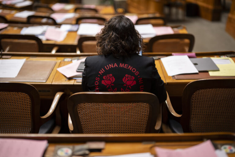 Vue de dos de Tamara Funiciello, assise dans un hémicycle au mobilier en bois, des rangées de bureaux jonchés de dossiers, de feuilles et d’un ordinateur portable devant elle, portant une veste noire ornée en rouge de l’inscription “Ni Una Menos” entourant deux roses stylisées.