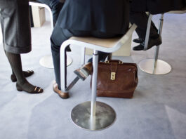 Bonnes nouvelles en novembre : Réduction des inégalités salariales, succès électoraux démocratiques et liaisons ferroviaires plus rapides en Europe Gros plan sur les jambes et les pieds de trois personnes en tenue professionnelle autour d’une table haute : deux assises sur des tabourets métalliques et une debout. Un porte-documents en cuir brun est posé sur la moquette bleu clair au pied de la table.