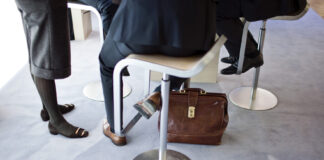 Bonnes nouvelles en novembre : Réduction des inégalités salariales, succès électoraux démocratiques et liaisons ferroviaires plus rapides en Europe Gros plan sur les jambes et les pieds de trois personnes en tenue professionnelle autour d’une table haute : deux assises sur des tabourets métalliques et une debout. Un porte-documents en cuir brun est posé sur la moquette bleu clair au pied de la table.