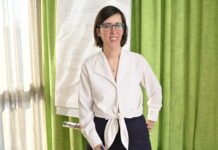 « Une femme en situation de handicap a 2 à 3 fois plus de risques d’être victime de violence » Céline Witschard, une femme souriante, vêtue d’un chemisier blanc noué à la taille et d’un pantalon sombre, porte des lunettes et tient un marqueur bleu. Elle se tient debout devant un paperboard sur lequel on distingue le mot « Accessibilité » et plusieurs questions (« Pour qui ? Comment ? Pourquoi ? »). En arrière-plan, un rideau vert et une fenêtre laissent filtrer la lumière du jour.