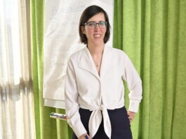 « Une femme en situation de handicap a 2 à 3 fois plus de risques d’être victime de violence » Céline Witschard, une femme souriante, vêtue d’un chemisier blanc noué à la taille et d’un pantalon sombre, porte des lunettes et tient un marqueur bleu. Elle se tient debout devant un paperboard sur lequel on distingue le mot « Accessibilité » et plusieurs questions (« Pour qui ? Comment ? Pourquoi ? »). En arrière-plan, un rideau vert et une fenêtre laissent filtrer la lumière du jour.