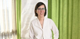 « Une femme en situation de handicap a 2 à 3 fois plus de risques d’être victime de violence » Céline Witschard, une femme souriante, vêtue d’un chemisier blanc noué à la taille et d’un pantalon sombre, porte des lunettes et tient un marqueur bleu. Elle se tient debout devant un paperboard sur lequel on distingue le mot « Accessibilité » et plusieurs questions (« Pour qui ? Comment ? Pourquoi ? »). En arrière-plan, un rideau vert et une fenêtre laissent filtrer la lumière du jour.