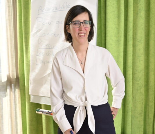 « Une femme en situation de handicap a 2 à 3 fois plus de risques d’être victime de violence » Céline Witschard, une femme souriante, vêtue d’un chemisier blanc noué à la taille et d’un pantalon sombre, porte des lunettes et tient un marqueur bleu. Elle se tient debout devant un paperboard sur lequel on distingue le mot « Accessibilité » et plusieurs questions (« Pour qui ? Comment ? Pourquoi ? »). En arrière-plan, un rideau vert et une fenêtre laissent filtrer la lumière du jour.