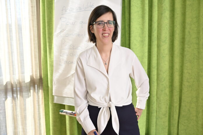 Céline Witschard, une femme souriante, vêtue d’un chemisier blanc noué à la taille et d’un pantalon sombre, porte des lunettes et tient un marqueur bleu. Elle se tient debout devant un paperboard sur lequel on distingue le mot « Accessibilité » et plusieurs questions (« Pour qui ? Comment ? Pourquoi ? »). En arrière-plan, un rideau vert et une fenêtre laissent filtrer la lumière du jour.