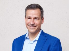 Les ONG sont sous pression : « Notre travail renforce la démocratie » Portrait d’un homme d’âge moyen, cheveux courts poivre et sel, souriant, vêtu d’une veste bleu vif et d’une chemise bleu clair. Il se tient debout, les mains dans les poches, devant un mur uni de couleur claire