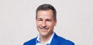 Les ONG sont sous pression : « Notre travail renforce la démocratie » Portrait d’un homme d’âge moyen, cheveux courts poivre et sel, souriant, vêtu d’une veste bleu vif et d’une chemise bleu clair. Il se tient debout, les mains dans les poches, devant un mur uni de couleur claire