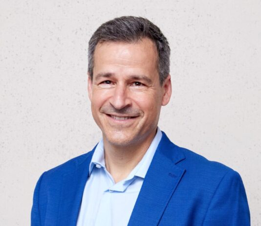 Les ONG sont sous pression : « Notre travail renforce la démocratie » Portrait d’un homme d’âge moyen, cheveux courts poivre et sel, souriant, vêtu d’une veste bleu vif et d’une chemise bleu clair. Il se tient debout, les mains dans les poches, devant un mur uni de couleur claire