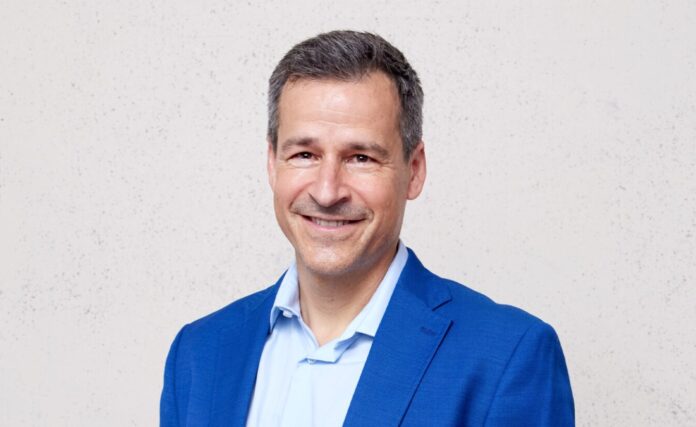 Felix_Gnehm_2 Portrait d’un homme d’âge moyen, cheveux courts poivre et sel, souriant, vêtu d’une veste bleu vif et d’une chemise bleu clair. Il se tient debout, les mains dans les poches, devant un mur uni de couleur claire