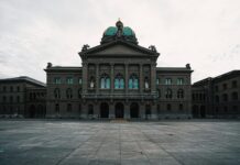 L’année politique 2026 en un coup d’œil Vue frontale du Palais fédéral à Berne, un vaste bâtiment néorenaissance en pierre grise bordé de colonnes et surmonté d’un dôme vert orné d’une statue. Au-dessus de l’entrée principale, l’inscription latine “Curia Confœderationis Helveticæ” se détache sur le fronton triangulaire. Devant la façade symétrique, la vaste place pavée—vide et légèrement humide sous un ciel nuageux—renforce l’impression de majesté solennelle.