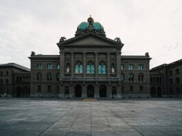 L’année politique 2026 en un coup d’œil Vue frontale du Palais fédéral à Berne, un vaste bâtiment néorenaissance en pierre grise bordé de colonnes et surmonté d’un dôme vert orné d’une statue. Au-dessus de l’entrée principale, l’inscription latine “Curia Confœderationis Helveticæ” se détache sur le fronton triangulaire. Devant la façade symétrique, la vaste place pavée—vide et légèrement humide sous un ciel nuageux—renforce l’impression de majesté solennelle.