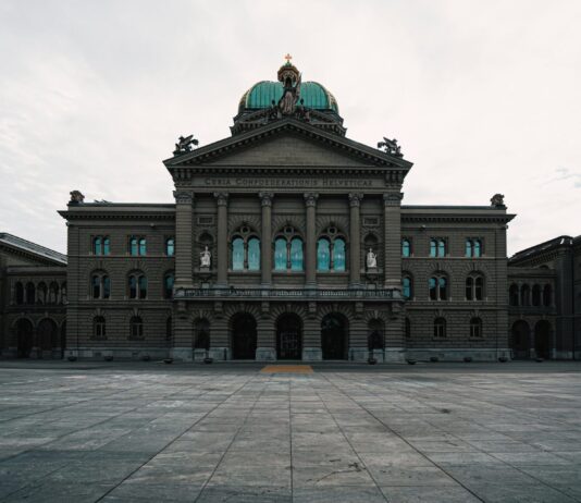 L’année politique 2026 en un coup d’œil Vue frontale du Palais fédéral à Berne, un vaste bâtiment néorenaissance en pierre grise bordé de colonnes et surmonté d’un dôme vert orné d’une statue. Au-dessus de l’entrée principale, l’inscription latine “Curia Confœderationis Helveticæ” se détache sur le fronton triangulaire. Devant la façade symétrique, la vaste place pavée—vide et légèrement humide sous un ciel nuageux—renforce l’impression de majesté solennelle.