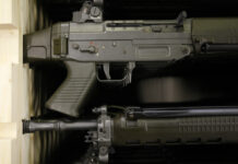 Affaiblissement de la loi sur le matériel de guerre : les exportations d’armes suisses vers des États autoritaires bientôt possibles Gros plan sur deux fusils d’assaut de couleur vert olive, rangés l’un au-dessus de l’autre dans une étagère en bois. On distingue la poignée pistolet, le boîtier de culasse avec le sélecteur de tir marqué “S” et chiffres rouges, la trappe d’éjection, la détente, ainsi que la partie avant du canon avec ses fixations et rails.