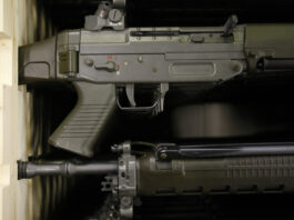 Affaiblissement de la loi sur le matériel de guerre : les exportations d’armes suisses vers des États autoritaires bientôt possibles Gros plan sur deux fusils d’assaut de couleur vert olive, rangés l’un au-dessus de l’autre dans une étagère en bois. On distingue la poignée pistolet, le boîtier de culasse avec le sélecteur de tir marqué “S” et chiffres rouges, la trappe d’éjection, la détente, ainsi que la partie avant du canon avec ses fixations et rails.