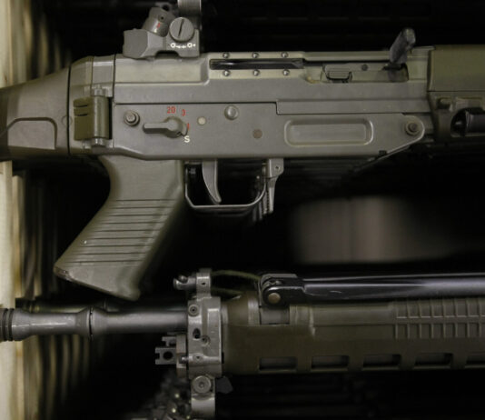 Affaiblissement de la loi sur le matériel de guerre : les exportations d’armes suisses vers des États autoritaires bientôt possibles Gros plan sur deux fusils d’assaut de couleur vert olive, rangés l’un au-dessus de l’autre dans une étagère en bois. On distingue la poignée pistolet, le boîtier de culasse avec le sélecteur de tir marqué “S” et chiffres rouges, la trappe d’éjection, la détente, ainsi que la partie avant du canon avec ses fixations et rails.