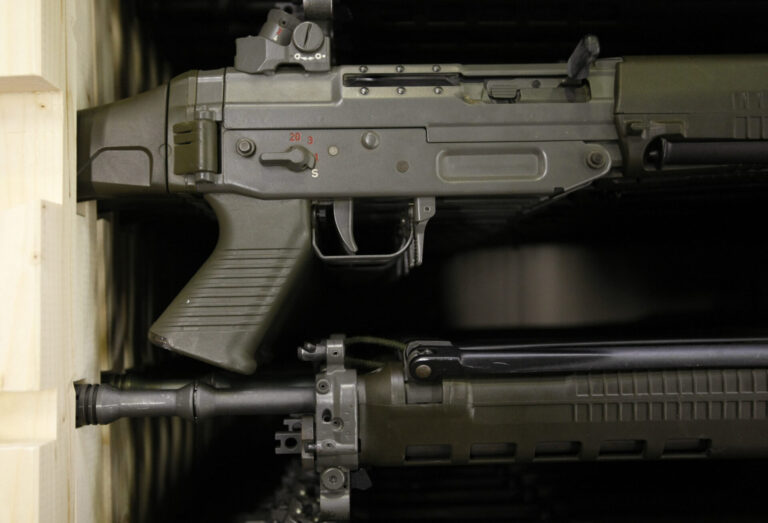 Gros plan sur deux fusils d’assaut de couleur vert olive, rangés l’un au-dessus de l’autre dans une étagère en bois. On distingue la poignée pistolet, le boîtier de culasse avec le sélecteur de tir marqué “S” et chiffres rouges, la trappe d’éjection, la détente, ainsi que la partie avant du canon avec ses fixations et rails.