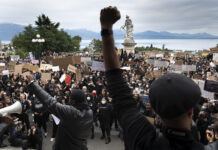 Bonnes nouvelles en décembre : lutte contre le racisme, plus de filles vont à l’école et revenu de base inconditionnel pour les Marshallais-es Une foule dense de manifestant·e·s se tient sur des marches et dans une esplanade, de dos et de face, brandissant de nombreuses pancartes (« Black Lives Matter », « No Justice No Peace », « La Suisse n’est pas innocente », etc.) et levant le poing en signe de solidarité. Au premier plan, un orateur de dos lève le poing, tandis qu’en arrière-plan on aperçoit une statue blanche et un lac bordé de montagnes sous un ciel nuageux.