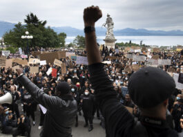 Bonnes nouvelles en décembre : lutte contre le racisme, plus de filles vont à l’école et revenu de base inconditionnel pour les Marshallais-es Une foule dense de manifestant·e·s se tient sur des marches et dans une esplanade, de dos et de face, brandissant de nombreuses pancartes (« Black Lives Matter », « No Justice No Peace », « La Suisse n’est pas innocente », etc.) et levant le poing en signe de solidarité. Au premier plan, un orateur de dos lève le poing, tandis qu’en arrière-plan on aperçoit une statue blanche et un lac bordé de montagnes sous un ciel nuageux.