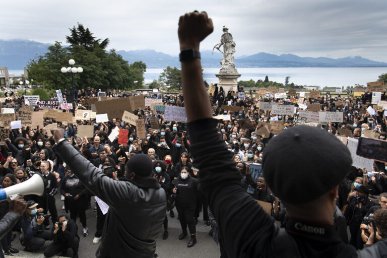 Une foule dense de manifestant·e·s se tient sur des marches et dans une esplanade, de dos et de face, brandissant de nombreuses pancartes (« Black Lives Matter », « No Justice No Peace », « La Suisse n’est pas innocente », etc.) et levant le poing en signe de solidarité. Au premier plan, un orateur de dos lève le poing, tandis qu’en arrière-plan on aperçoit une statue blanche et un lac bordé de montagnes sous un ciel nuageux.