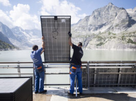 Initiative pour un fonds climat : investir dans une Suisse sûre et agréable à vivre Deux techniciens en tenue de travail bleue soulèvent et positionnent un grand panneau solaire contre une rambarde métallique, avec derrière eux un vaste réservoir d’eau d’un vert pâle encerclé de falaises et de sommets rocheux sous un ciel parsemé de nuages.