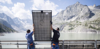 Initiative pour un fonds climat : investir dans une Suisse sûre et agréable à vivre Deux techniciens en tenue de travail bleue soulèvent et positionnent un grand panneau solaire contre une rambarde métallique, avec derrière eux un vaste réservoir d’eau d’un vert pâle encerclé de falaises et de sommets rocheux sous un ciel parsemé de nuages.