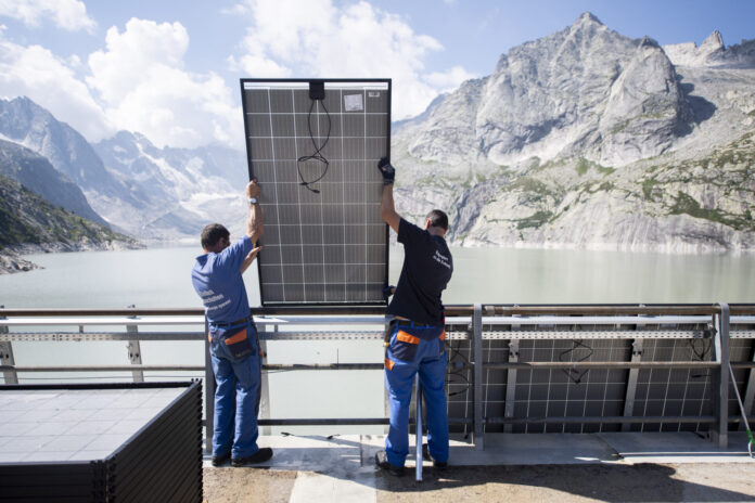 SOLARANLAGE, STAUSEE, STAUDAMM, STAUMAUER, ALBIGNA, SOLARMODULE, SOLAR MODULE, SOLARZELLEN, SOLARZELLE, SOLARZELLEN, Deux techniciens en tenue de travail bleue soulèvent et positionnent un grand panneau solaire contre une rambarde métallique, avec derrière eux un vaste réservoir d’eau d’un vert pâle encerclé de falaises et de sommets rocheux sous un ciel parsemé de nuages.