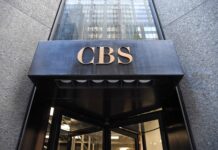 Comment des milliardaires ont transformé une chaîne de télévision américaine traditionnelle en une machine de propagande Entrée d’un immeuble : au-dessus d’une porte tournante en verre, un auvent en métal noir supporte en son centre le sigle « CBS » en lettres dorées. La façade autour est en pierre grise.