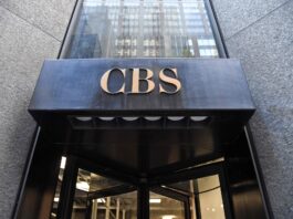 Comment des milliardaires ont transformé une chaîne de télévision américaine traditionnelle en une machine de propagande Entrée d’un immeuble : au-dessus d’une porte tournante en verre, un auvent en métal noir supporte en son centre le sigle « CBS » en lettres dorées. La façade autour est en pierre grise.