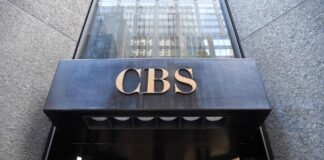 Comment des milliardaires ont transformé une chaîne de télévision américaine traditionnelle en une machine de propagande Entrée d’un immeuble : au-dessus d’une porte tournante en verre, un auvent en métal noir supporte en son centre le sigle « CBS » en lettres dorées. La façade autour est en pierre grise.