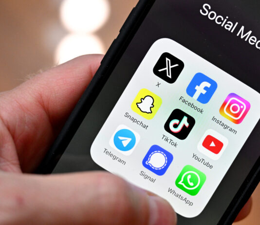 Initiative anti-SSR : plus de fausses nouvelles, moins de vérification Gros plan sur une main tenant un smartphone : à l’écran, un dossier nommé « Social Media » affiche neuf icônes d’applications disposées en grille : X (anciennement Twitter), Facebook, Instagram, Snapchat, TikTok, YouTube, Telegram, Signal et WhatsApp. L’arrière-plan est flou, mettant l’accent sur l’écran du téléphone.