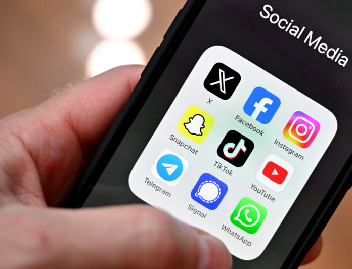 Gros plan sur une main tenant un smartphone : à l’écran, un dossier nommé « Social Media » affiche neuf icônes d’applications disposées en grille : X (anciennement Twitter), Facebook, Instagram, Snapchat, TikTok, YouTube, Telegram, Signal et WhatsApp. L’arrière-plan est flou, mettant l’accent sur l’écran du téléphone.