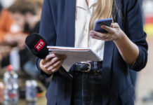 L’information en Suisse romande est menacée par l’initiative anti-SSR Gros plan sur une journaliste en tenue professionnelle (blazer bleu marine et chemisier blanc) : dans sa main gauche, elle tient un micro noir marqué « RTS » et un petit calepin à spirales, dans sa main droite, un smartphone à coque bleue. L’arrière-plan, flou, suggère un environnement intérieur animé.