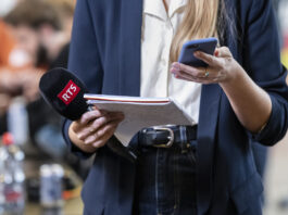 L’information en Suisse romande est menacée par l’initiative anti-SSR Gros plan sur une journaliste en tenue professionnelle (blazer bleu marine et chemisier blanc) : dans sa main gauche, elle tient un micro noir marqué « RTS » et un petit calepin à spirales, dans sa main droite, un smartphone à coque bleue. L’arrière-plan, flou, suggère un environnement intérieur animé.