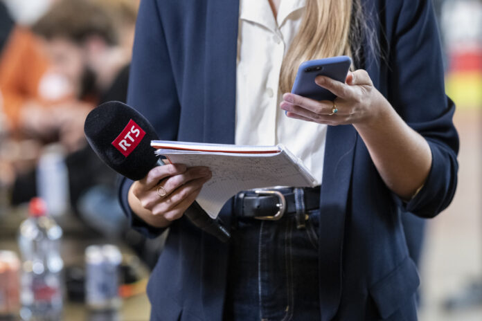 Gros plan sur une journaliste en tenue professionnelle (blazer bleu marine et chemisier blanc) : dans sa main gauche, elle tient un micro noir marqué « RTS » et un petit calepin à spirales, dans sa main droite, un smartphone à coque bleue. L’arrière-plan, flou, suggère un environnement intérieur animé.