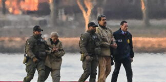 Venezuela : Trump mise illégalement sur « le droit du plus fort » « Sur un quai en bord d’eau, quatre agents en tenue tactique — dont certains portent des blousons marqués “DEA” — escortent Nicolàs Maduro et Cilia Flores menottées vêtues de blousons et pantalons beiges. Le soleil couchant teinte le ciel d’orangé, et des blocs de béton peints en rouge et blanc délimitent le quai devant une surface d’eau calme. »