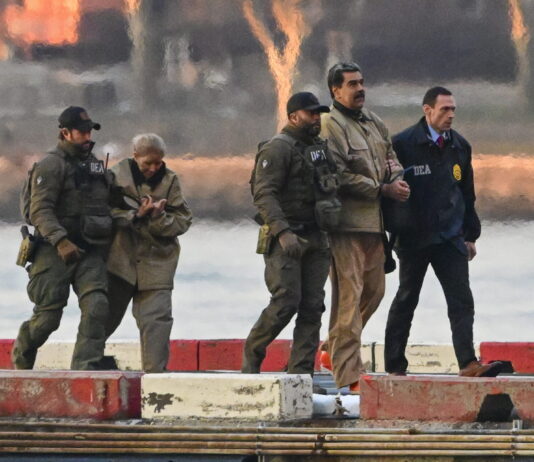 Venezuela : Trump mise illégalement sur « le droit du plus fort » « Sur un quai en bord d’eau, quatre agents en tenue tactique — dont certains portent des blousons marqués “DEA” — escortent Nicolàs Maduro et Cilia Flores menottées vêtues de blousons et pantalons beiges. Le soleil couchant teinte le ciel d’orangé, et des blocs de béton peints en rouge et blanc délimitent le quai devant une surface d’eau calme. »